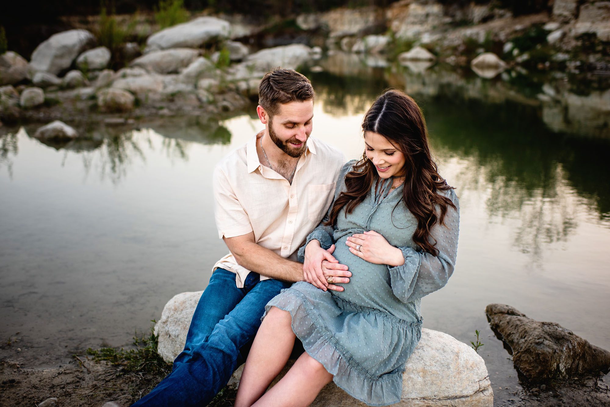 Couple sitting on a rock near a lake looking at mom's baby bump, Maternity photographer in San Antonio