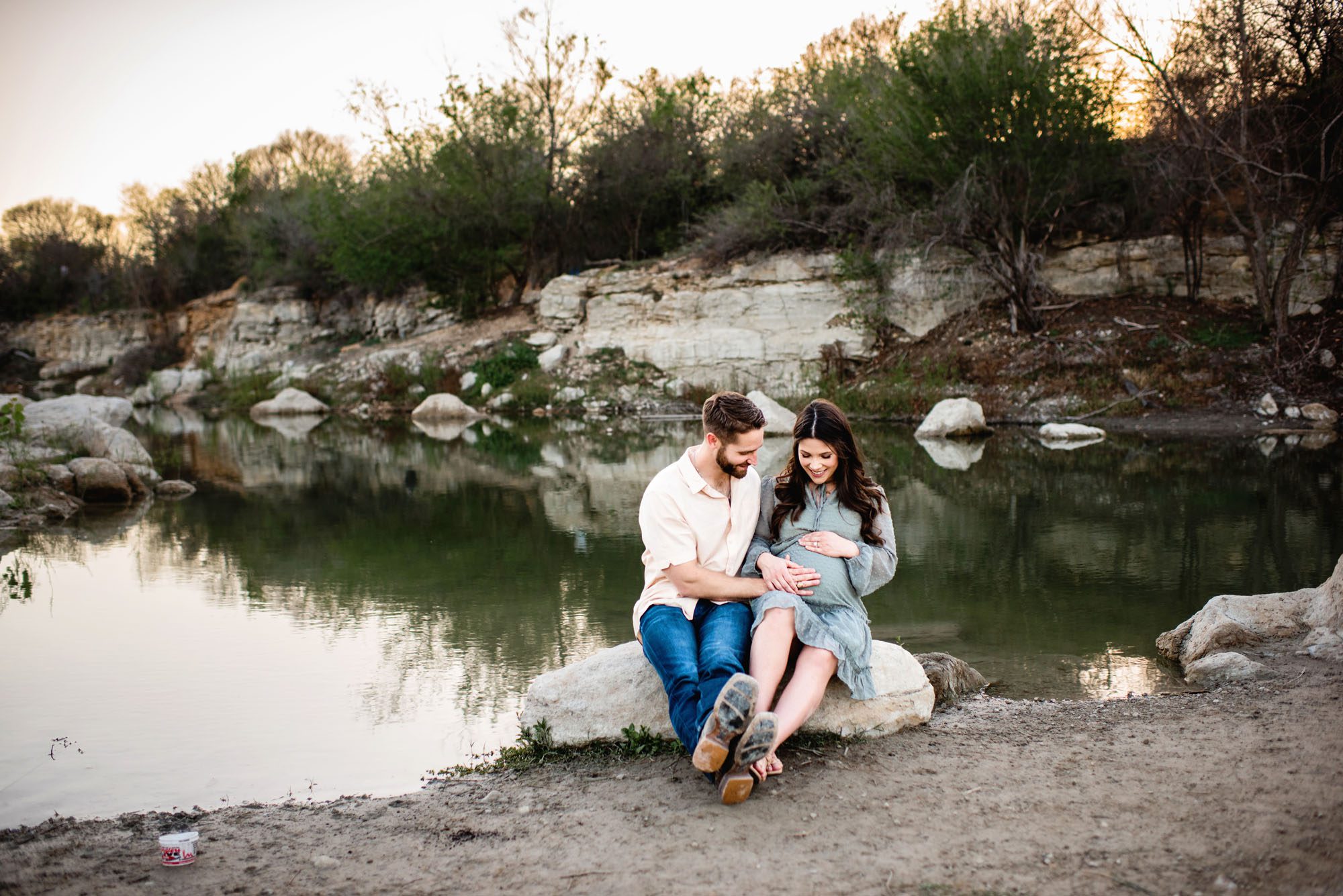 Couple sitting on a rock near a lake looking at mom's pregnant belly, San Antonio maternity photographer