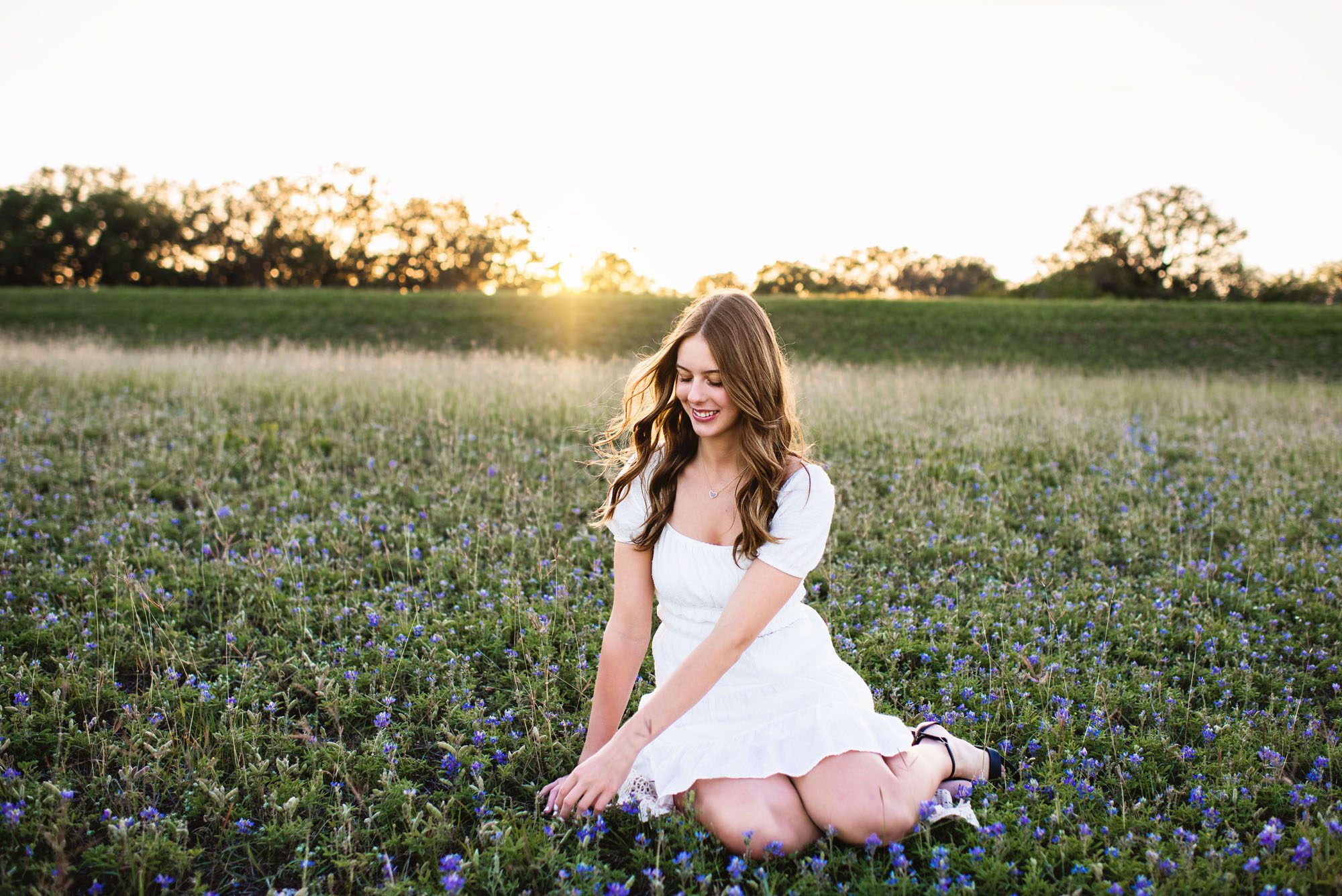Girl in white dress sitting in bluebonnet field touching flowers, San Antonio senior photography