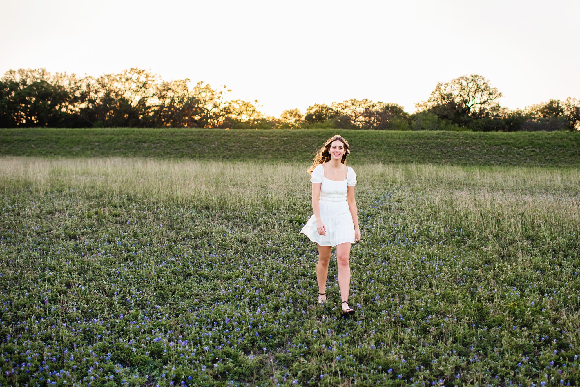 Senior in white dress walking in bluebonnet field, San Antonio senior photographer
