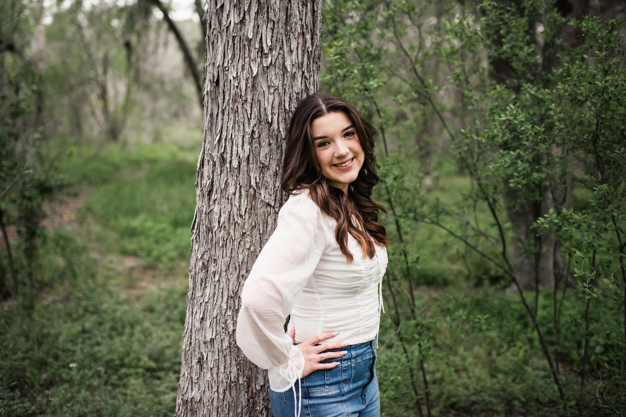 Girl in white shirt and jeans standing by tree smiling at camera, San Antonio senior photographer