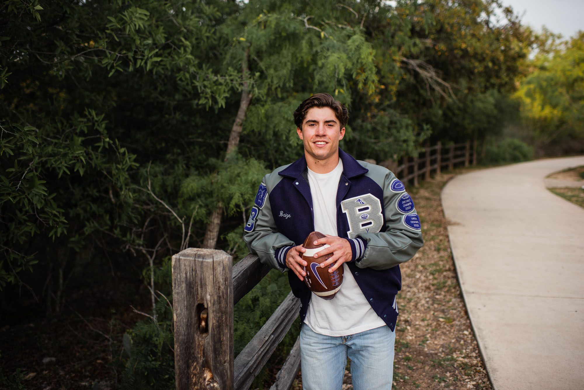 Boy holding football with letter jacket leaning on wooden fence, San Antonio senior photographer