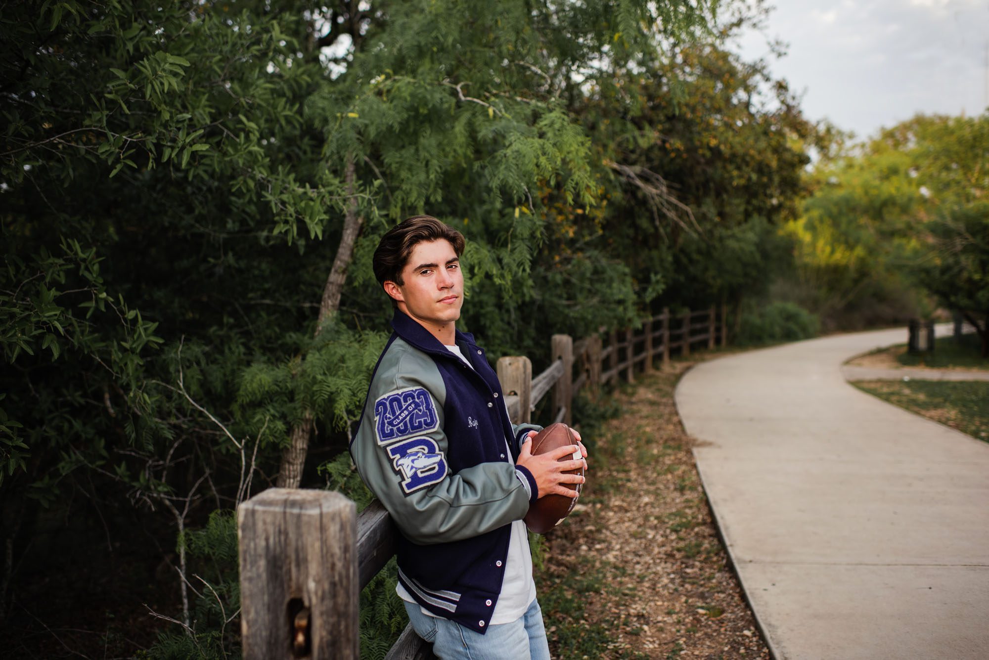 Boy holding football with letter jacket by wooden fence, San Antonio senior photographer