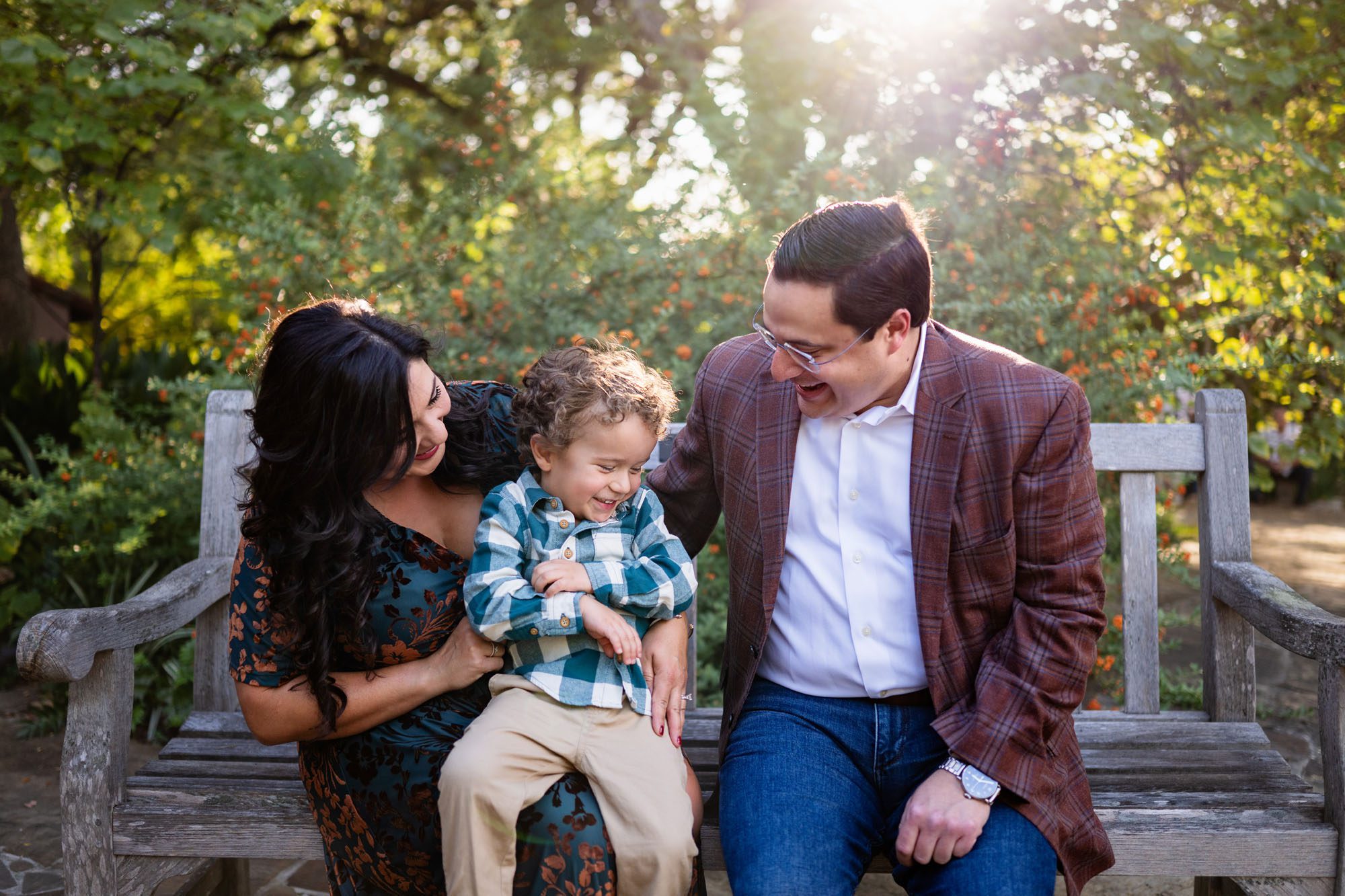 Mom and dad tickling son on bench with glowing sun behind them, San Antonio family photography